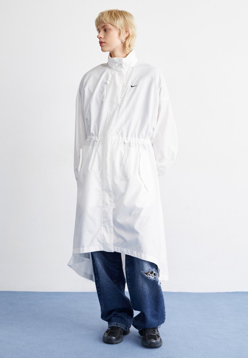 Witte lange regenjas met een hoge kraag en verstelbare taille, voorzien van een klein zwart logo, gecombineerd met losse blauwe jeans en zwarte schoenen.
