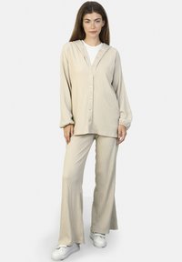 Beiges, strukturiertes Loungewear-Set mit einer Kapuzenjacke mit Kn�öpfen und langen Ärmeln sowie passenden, schmal zulaufenden Hosen. Getragen mit weißen Sneakern.