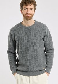 Gilet en tricot gris avec un motif texturé, encolure ronde et manches et ourlet côtelés. Petit logo en accent sur le côté inférieur.