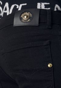 Jeans de mezclilla negros con un tejido texturizado, herrajes en tono dorado y un parche de cuero con marca que presenta un emblema circular en la cintura.