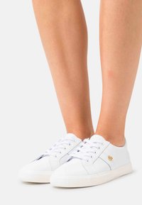 White leather sneakers with laces and a small gold logo worn on bare feet against a plain background.