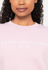 Világos rózsaszín pulóver bordázott nyakkivágással és fehér nyomtatott szöveggel: "TOMMY HILFIGER EST. 1985" az elején. Simább, laza anyag.