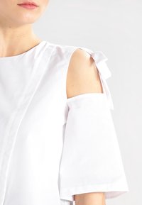 Biała bluzka z krótkimi rękawami, z wyciętymi ramionami i akcentami w postaci kokardek. Wykonana z gładkiego materiału w czystym, minimalistycznym designie.