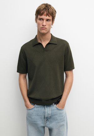 Jonge man met lichtbruin haar, gekleed in een donkergroen polo met korte mouwen en lichtblauwe spijkerbroek, staand tegen een effen witte achtergrond.
