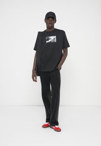 Μαύρο μπλουζάκι με φωτογραφικό σχέδιο, σε συνδυασμό με σκούρο γκρι παντελόνι και κόκκινα αθλητικά παπούτσια. Μια μαύρη καπέλο ολοκληρώνει την εμφάνιση, προσδίδοντας σπορτίφ χαρακτήρα.