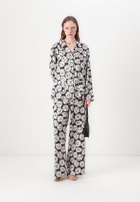 Kukkakuvioinen pyjamapuku mustana, jossa on suuria valkoisia päivänkakkaroita oransseilla keskustoilla. Löysät pitkät hihat ja leveälahkeiset housut, yhdistettynä mustaan laukkuun.