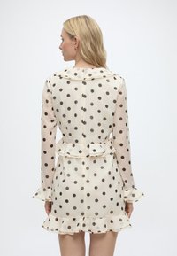 Ξανθιά γυναίκα γυρισμένη με την πλάτη φοράει κρέμ φόρεμα με μαύρες πουά, με βολάν στα μανίκια, στο τελείωμα, στη μέση και με γιακά.