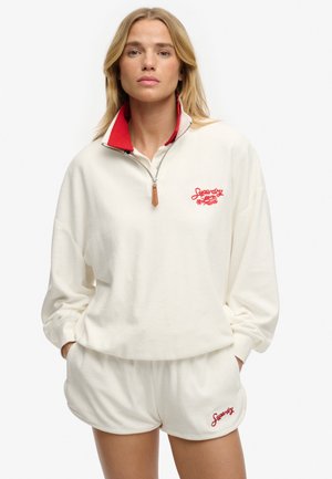 Kobieta ubrana w biały zestaw dresowy z suwakiem i krótkie spodenki z czerwonymi haftowanymi logo "Superdry", stojąca z rękami w kieszeniach na białym tle.