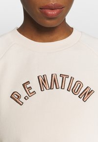 Ljusbeige sweatshirt med ribbad rundhals och broderad text "P.E NATION" i svart och koppar. Har raglanärmar och en mjuk tygtextur.
