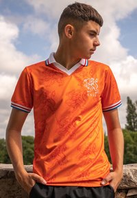 Oranje voetbalshirt met een leeuwenpatroon, voorzien van een witte kraag en blauwe strepen op de mouwen. Lichtgewicht, atletische pasvorm.