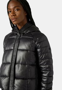 Giacca a piumino nera con finitura lucida, cuciture orizzontali, cappuccio con coulisse e patta anteriore dalla texture lavorata.