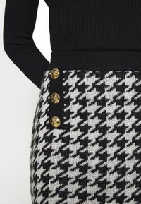 Jupe à motif pied-de-poule noir et blanc avec trois boutons dorés sur le côté, portée avec un haut noir à manches longues côtelé.