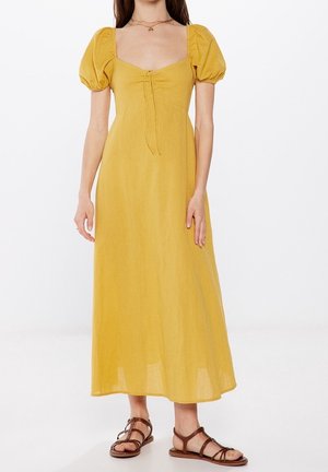 Mujer con un vestido midi amarillo mostaza con mangas abullonadas, escote cuadrado, detalle de lazo en el frente y sandalias planas marrones con tiras.