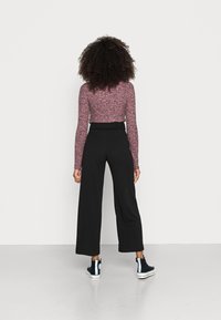 Vrouw met krullend haar die een lang mouwige roze gemêleerde top, hoge zwarte wijde broek en zwarte hoge sneakers draagt, staand met de rug naar ons toegekeerd.