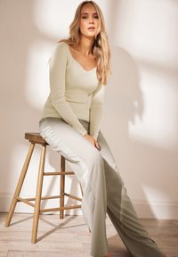 Haut à col en V à manches longues, de couleur vert clair et côtelé, associé à un pantalon large gris clair avec des accents en satin sur les côtés. Assise sur un tabouret en bois.