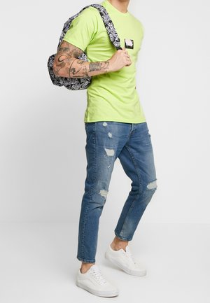 T-shirt in cotone verde neon con una piccola patch stampata, abbinato a jeans in denim blu strappati. Sneakers bianche e uno zaino con motivo in bianco e nero.