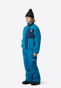 Μπλε φλις μπουφάν με ναυτικά στοιχεία, τσέπες με φερμουάρ, αντίστοιχο μπλε παντελόνι και μαύρο χειμερινό καπέλο με λευκό σχέδιο. Μαύρα παπούτσια.