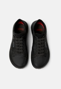 Schwarze Sportschuhe mit einem Obermaterial aus Mesh, flachen Schnürsenkeln, rotem Innenfutter und flexibler Gummisohle. Mit "CAMPER"-Branding auf der Zunge und der Seite.