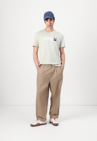 Light green short-sleeve t-shirt with "TOMMY" printed in white and red. Beige wide-leg trousers, white sneakers with brown accents, and a blue cap.