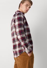 Langärmliges Karhemd in Burgunderrot, Weiß und Marineblau mit Knopfmanschetten; kombiniert mit strukturierten braunen Cordhosen.