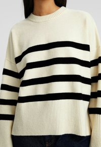 Maglione color crema con righe orizzontali nere, colletto e orlo a costine, realizzato in materiale morbido, con maniche a taglio morbido e una forma rilassata.
