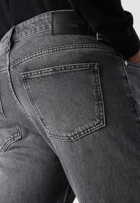 Grå denimbukser med middels høyde, utstyret med baklommer og et lærmerke i midjen. Teksturlignende stoff med sømdetaljer.
