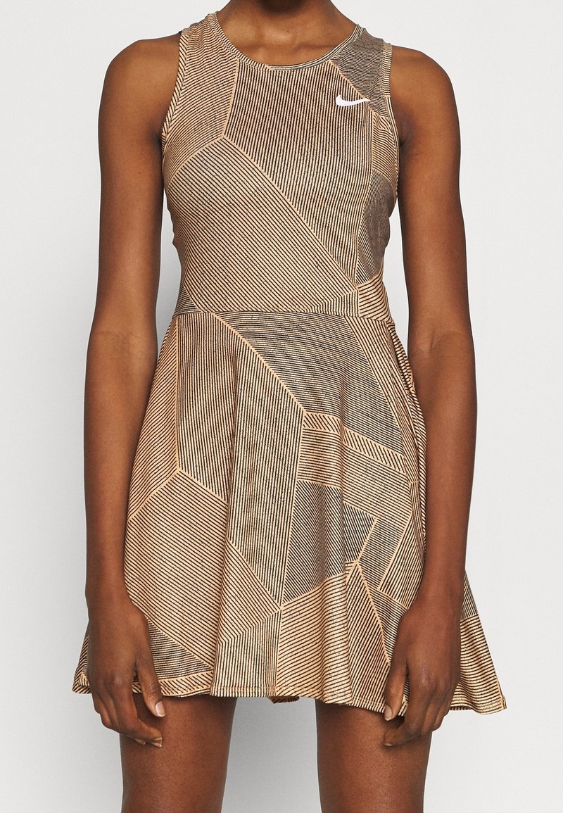 Femme portant une robe sans manches beige et noire à motif géométrique, jupe évasée et petit logo blanc sur la poitrine.