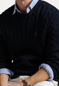 Pull en maille torsadée marine avec un col rond et un petit logo rouge, superposé à une chemise à col rayée bleu et blanc.