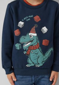 Tumši zila sporta krekla augšdaļa ar karikatūras T-Rexu, kas valkā Ziemassvētku cepuri un šalli, apkārt ir krāsaini dāvanas. Mīksta auduma, ribota apkakle.