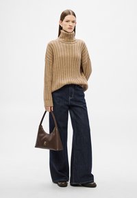 Μπεζ ριμπ πουλόβερ με ζιβάγκο, φαρδιά σκούρα τζιν παντελόνια και καφέ δερμάτινη τσάντα με χρυσά μεταλλικά στοιχεία. Το μοντέλο στέκεται μπροστά σε λευκό φόντο.