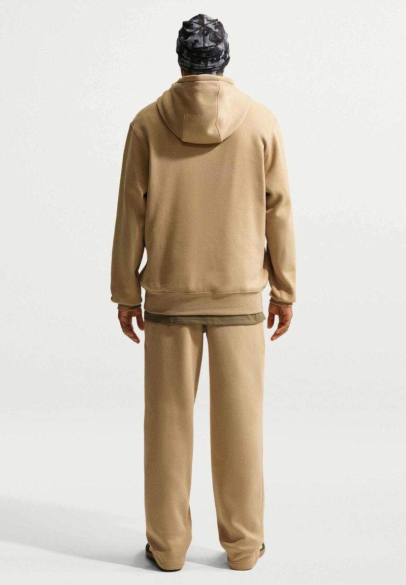 Mann trägt beige Kapuzenpullover und passende Hose, Mütze mit Tarnmuster, steht mit dem Rücken zur Kamera vor hellem einfarbigem Hintergrund.