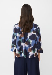 Donna con lunghi capelli castani che indossa una camicia larga con un motivo astratto multicolore e pantaloni neri ampi, in piedi di spalle contro uno sfondo bianco.