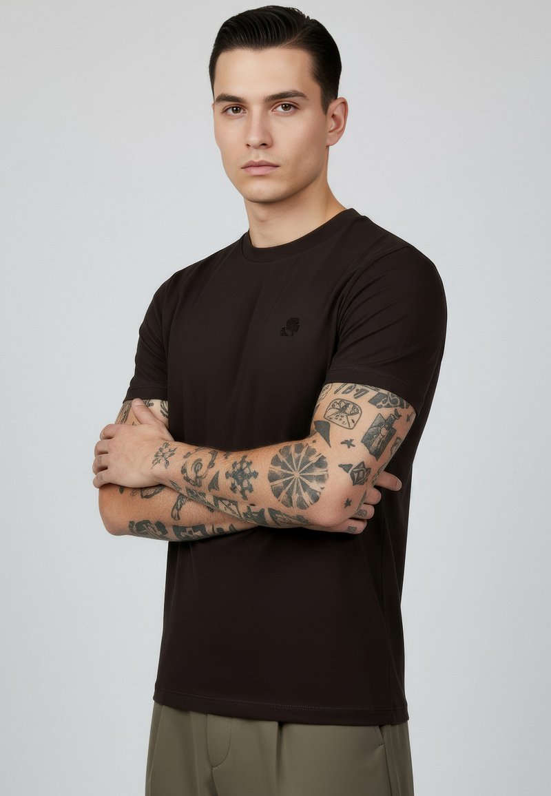 Jeune homme aux cheveux lissés en arrière portant un t-shirt noir, bras croisés, montrant des tatouages noirs détaillés sur les deux avant-bras.