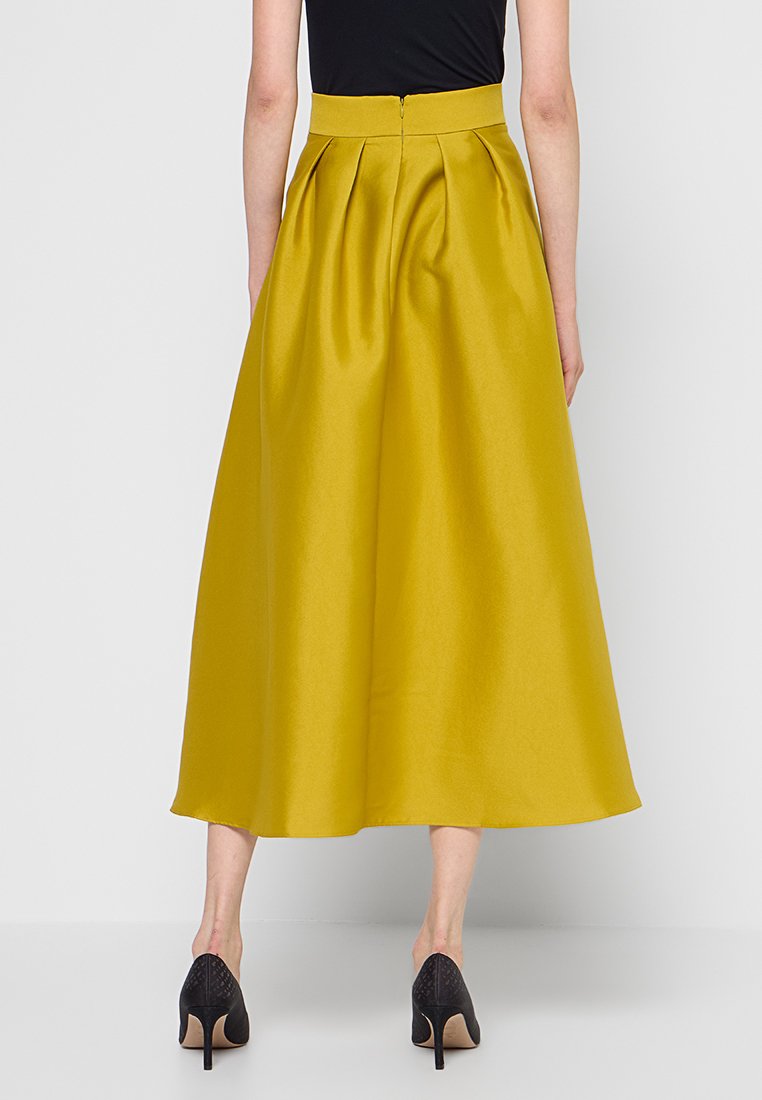 Plissé, middellange rok van vibrerend gele satijnen stof, met een hoge taille en een verborgen rits aan de achterkant, gecombineerd met zwarte hoge hakken.
