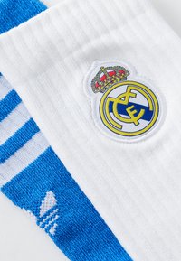 Hvide fodboldstrømper med et blåt og hvidt stribet mønster, prydet med et broderet Real Madrid-logo og krone-detalje.