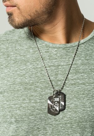 Homme barbu portant une chemise verte chinée et un collier en chaîne argentée avec deux pendentifs noir Diesel en forme de plaques militaires.