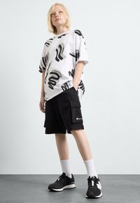 Hvit grafisk t-skjorte med svarte abstrakte mønstre, svarte cargo-shorts med sidelommer, hvite sokker og svarte sportssko med hvit såle.
