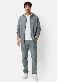 Lichtgrijze hoodie met ritssluiting en twee zakken aan de voorkant, gecombineerd met lichtgroene cargobroek met zijzakken en witte sneakers.