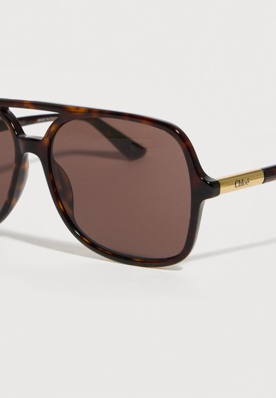 Γυαλιά ηλίου τετράγωνου σχήματος με καβούκι χελώνας, φακούς με καφέ απόχρωση και χρυσά μπράτσα χαραγμένα με το λογότυπο «Chloé» σε λευκό φόντο.