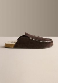 Chausson homme en daim brun foncé à enfiler avec coutures blanches et doublure beige douce, semelle plate, vu de côté sur une surface neutre.