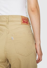 Béžové džínové kalhoty s vysokým pasem, s červenou koženou značkovou nášivkou a charakteristickým prošíváním zadní kapsy ve tvaru srdce.