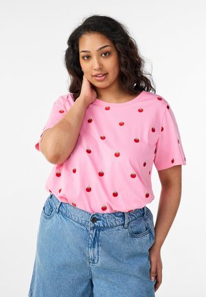 Kvinde med bølget mørkt hår iført en pink T-shirt med tomatmønster og højttaljede blå jeans, poserer med den ene hånd på halsen.