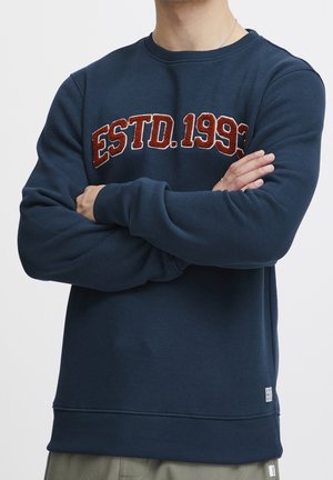 Homme portant un sweat-shirt bleu marine avec l'inscription « ESTD. 1993 » en lettres rouges texturées, les bras croisés, et un collier en chaîne argentée visible.