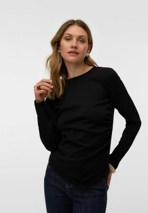 Femme aux cheveux châtain clair portant des boucles d'oreilles argentées et une chemise noire à manches longues, posant avec une main près du visage et l'autre sur la hanche.