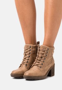 Botas de tobillo de ante color crema con cordones y cremallera lateral, con tacones de bloque, llevadas sobre piernas desnudas contra un fondo blanco.