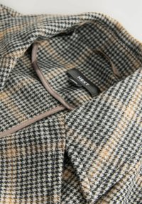 Kragen und Ausschnitt eines karierten Wollmantels in Schwarz, Weiß und Braun, mit sichtbarem NEXT-Label innen.