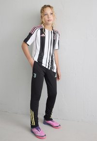 Jersey de Adidas a rayas blancas y negras con acentos rosas, pantalones negros con logo y rayas amarillas, y botas de fútbol coloridas.