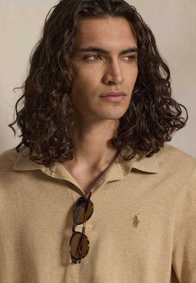 Νέος άνδρας με μπούκλες μέχρι τους ώμους, που φοράει μπεζ πουκάμισο με γιακά και γυαλιά ηλίου κρεμασμένα στο λαιμό.