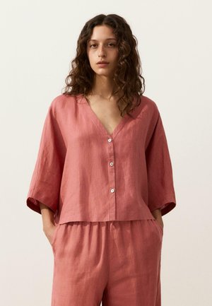 Młoda kobieta o kręconych włosach, ubrana w luźną lnianą bluzkę na guziki w kolorze przygaszonego różu oraz dopasowane spodnie, stojąca z rękami w kieszeniach na jednolitym tle.