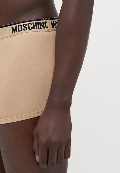 Beige boxer nadrág fekete Moschino logóval díszített derékrészel, sima textúrával és testhezálló fazonnal, oldalnézetből megörökítve.